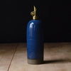 A tall cylindrical blue ceramic oil bottle with a glossy speckled glaze, a dark gray base, and a brass-toned decorative spout or cap, standing on a light wood surface against a dark background.