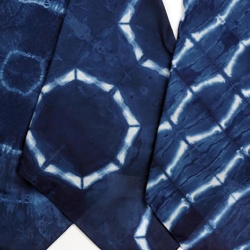 A close-up of dark indigo blue fabric with a white shibori tie-dye pattern, showing geometric hexagonal and grid-like markings.