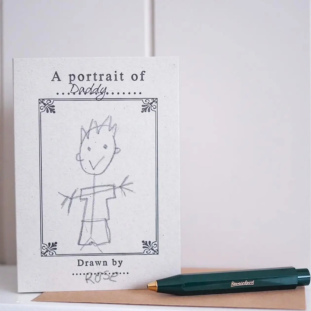 A square white card or print with the text 'A portrait of Daddy' and a simple hand-drawn sketch of a child, placed on a tan surface next to a dark green pen with gold lettering.