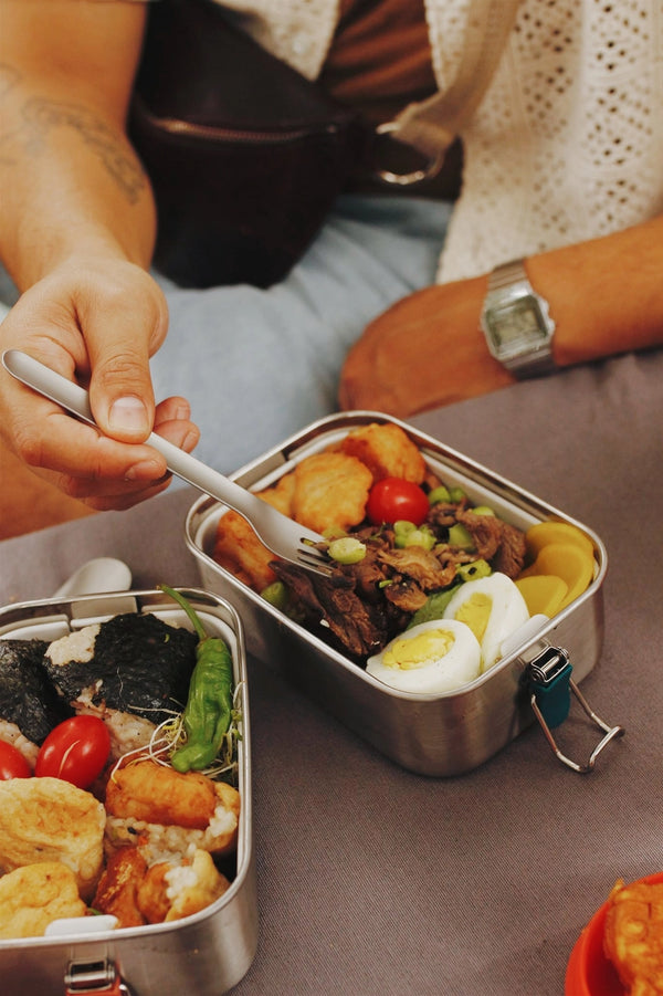 Person eating from a metal bento box filled with food on a table.