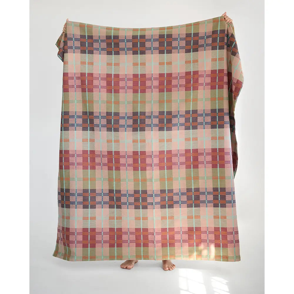 A large picnic blanket is held up by a person whose bare feet are visible at the bottom, against a plain light gray studio background.