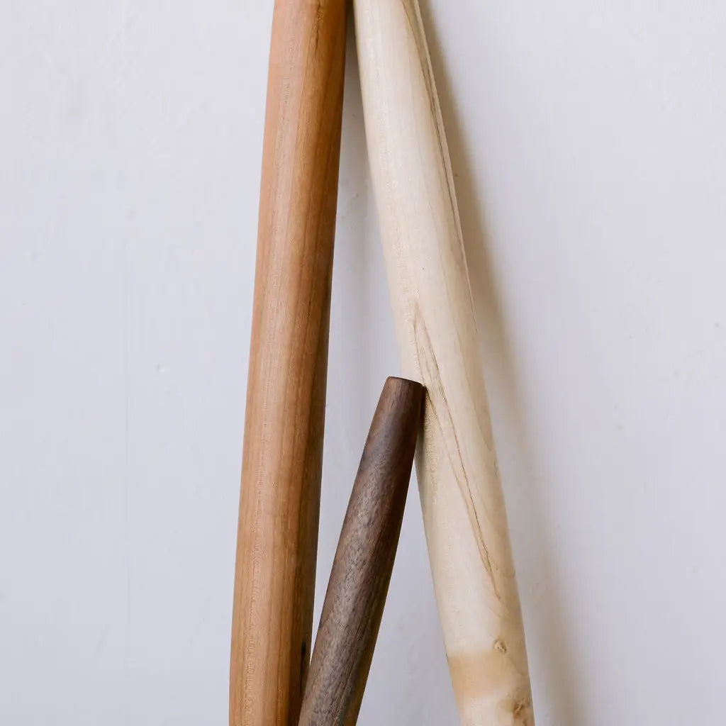 Three wooden rolling pins in natural light brown, cream, and dark brown tones, arranged vertically against a plain off-white background.