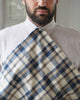 A close-up of a man wearing a light-colored linen shirt with a blue, beige, and brown plaid scarf draped over his chest.