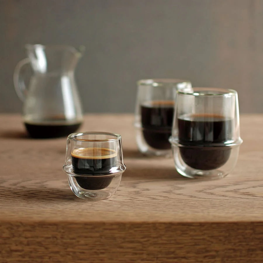 Three small glass cups with espresso on a wooden surface, with a pitcher in the background.