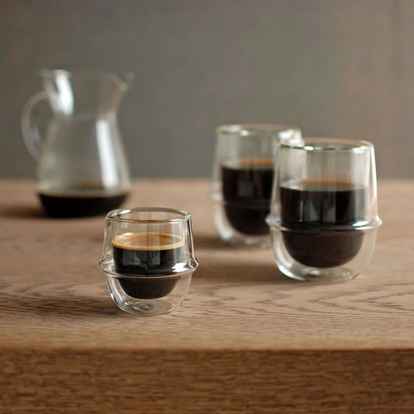 Three small glass cups with espresso on a wooden surface, with a pitcher in the background.