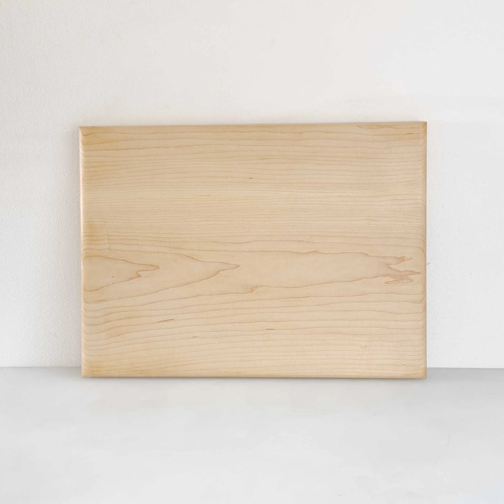 A rectangular light wood cutting board with visible natural grain patterns, standing upright against a plain white wall on a light gray surface.