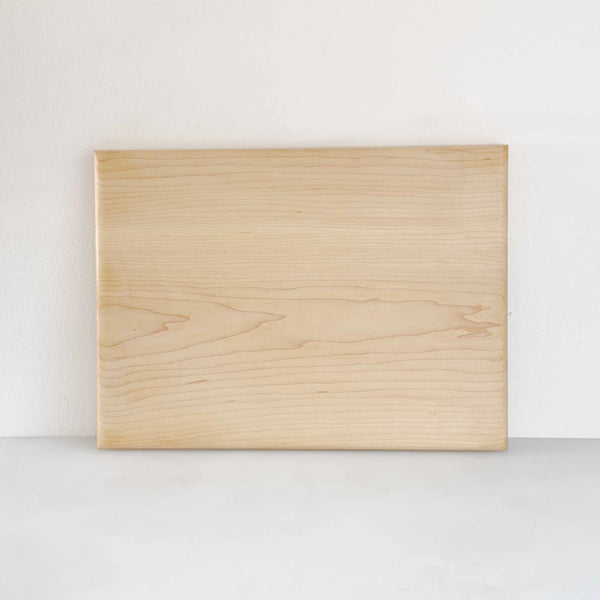 A rectangular light wood cutting board with visible natural grain patterns, standing upright against a plain white wall on a light gray surface.