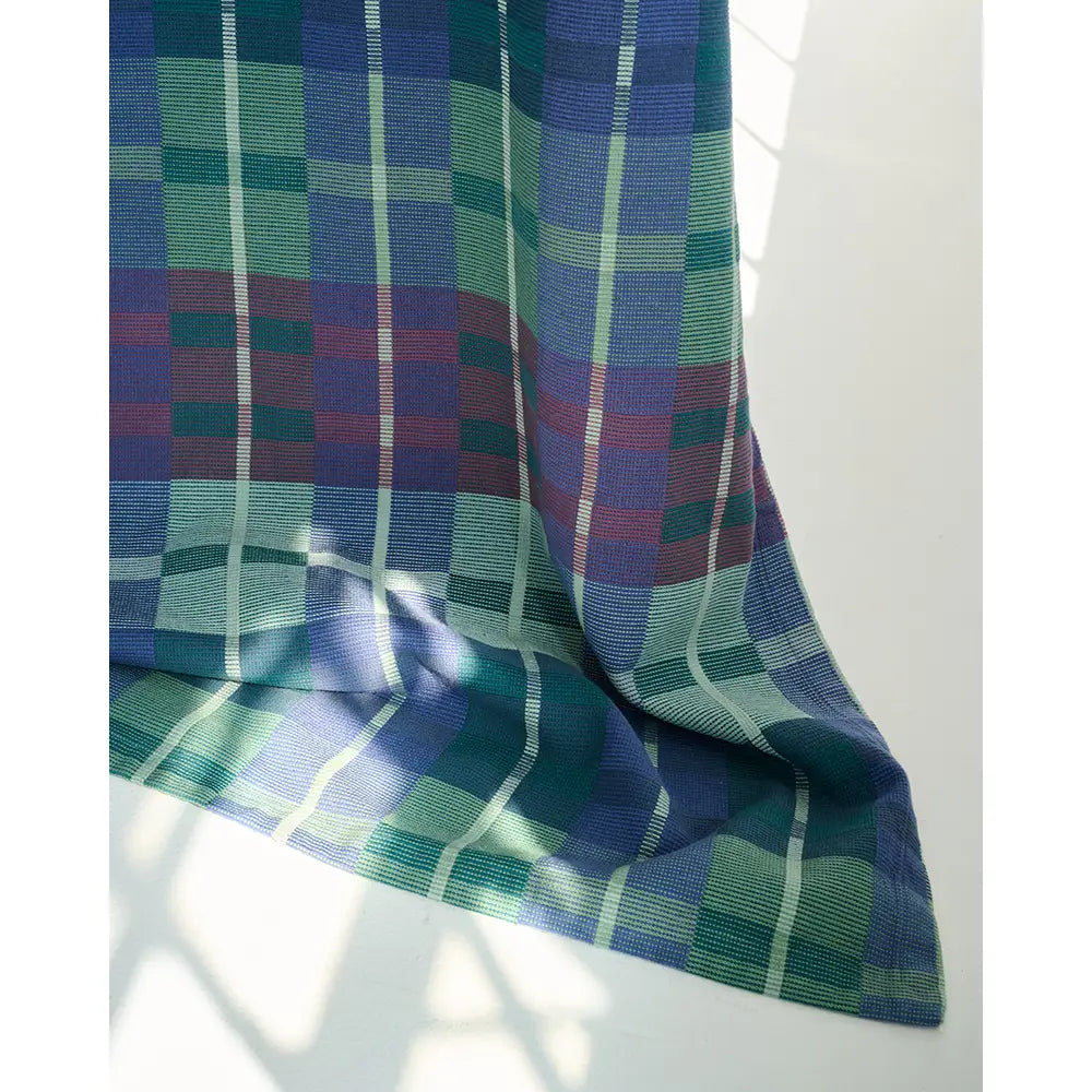 A picnic blanket, featuring a tartan pattern in shades of green, blue, purple, and white, draped against a white background with soft window light shadows.