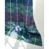 A picnic blanket, featuring a tartan pattern in shades of green, blue, purple, and white, draped against a white background with soft window light shadows.