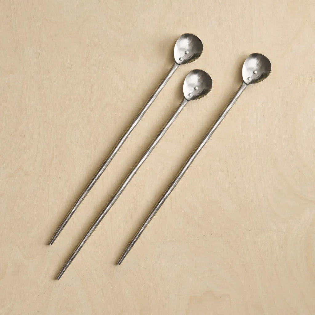 Three long metal stirring spoons with hammered oval heads, arranged diagonally on a light beige wood background.