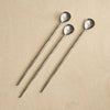 Three long metal stirring spoons with hammered oval heads, arranged diagonally on a light beige wood background.