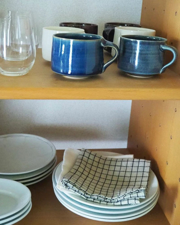 Set of blue mugs, clear glasses, and white plates with a checkered towel on a wooden shelf.