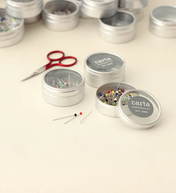 Silver tins with pins and a red scissors on a light background