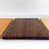 Three rectangular wooden cutting boards with rounded corners displayed side-by-side, featuring different wood grain patterns and tones: a dark brown walnut board in the center, flanked by two lighter natural wood boards on either side, set against a plain white background.