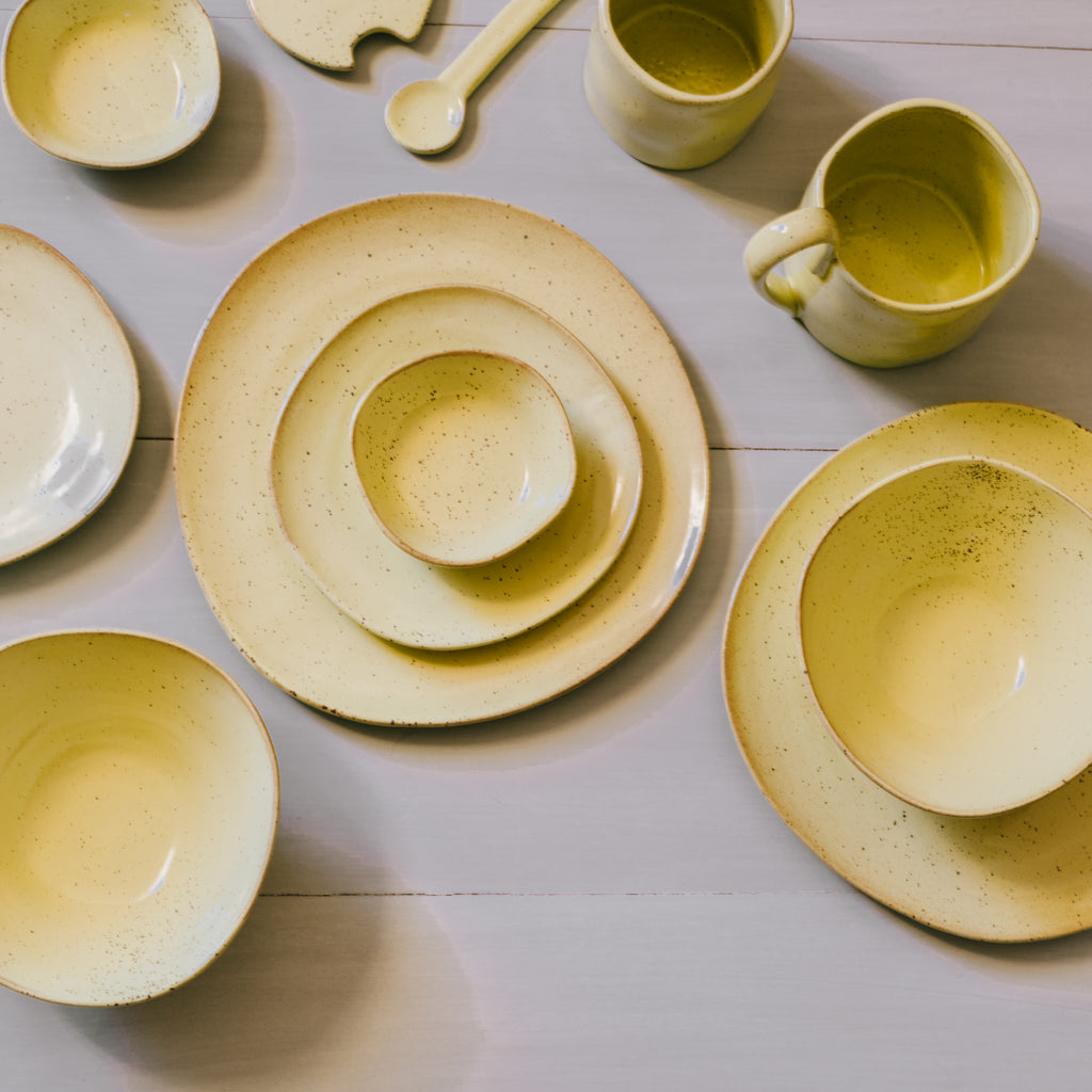 A collection of porcelain plates in various sizes and a small ceramic cup, all with a semi-matte yellow glaze, displayed on a wooden surface.