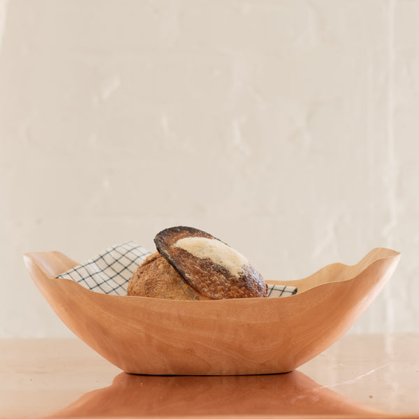 Wooden oval mango bowl with bread and a checkered tea towel inside