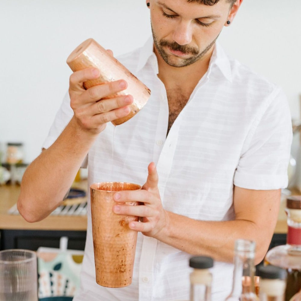 A person holding a copper cocktail shaker with a lid, in a kitchen setting with various tools and ingredients in the background.